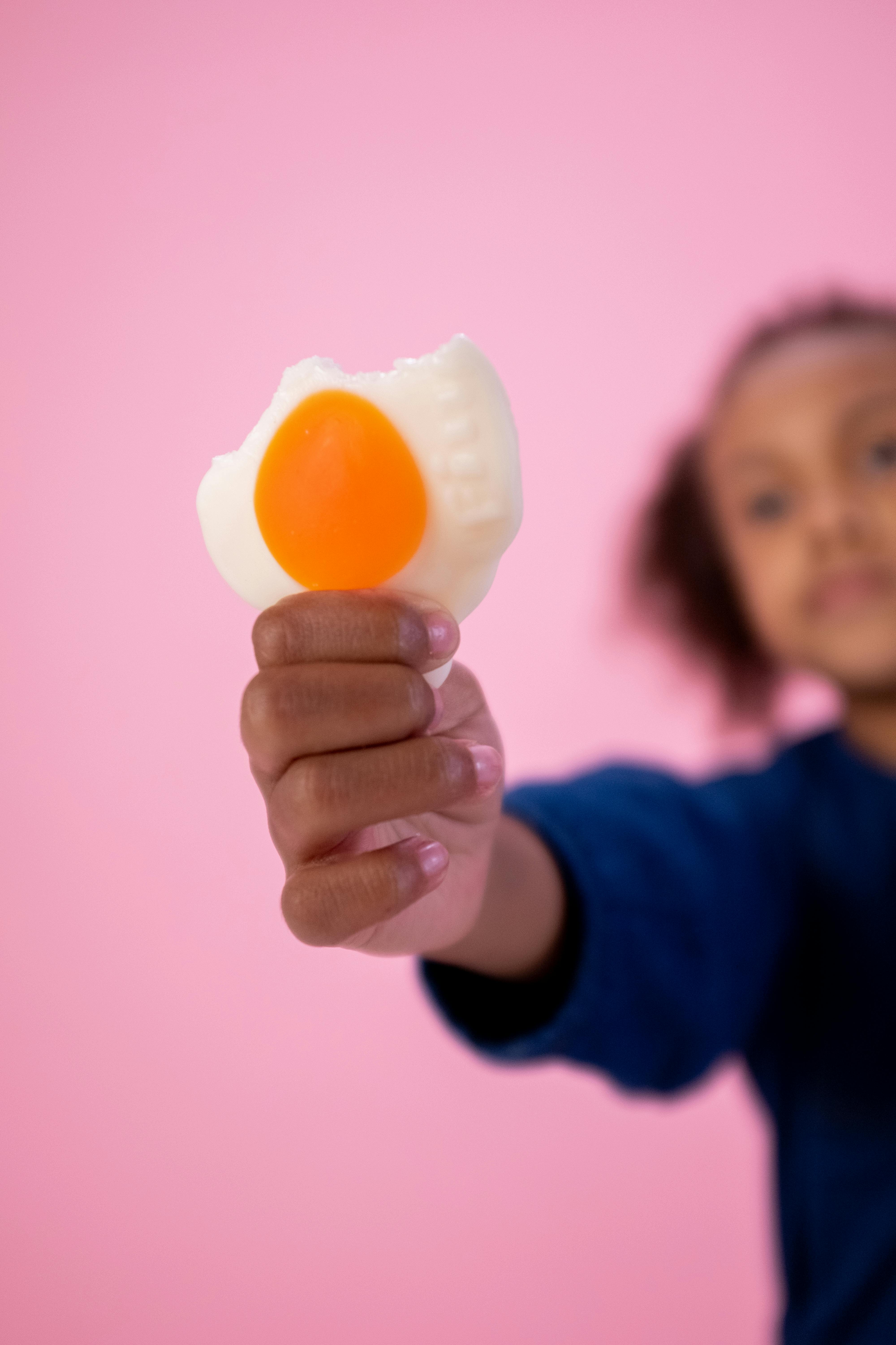 A child holds a bitten gummy candy against a pink studio background, expressing a sense of playfulness and sweetness.