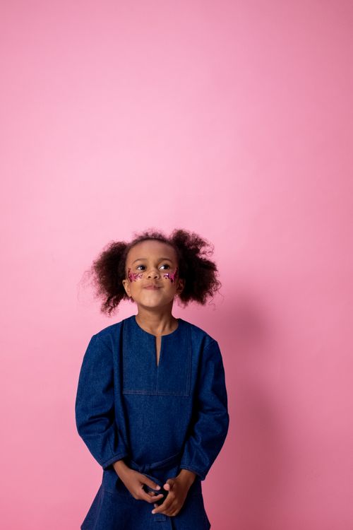 Free Adorable child with face stickers, in a denim dress, stands happily against a pink background. Perfect for playful themes. Stock Photo