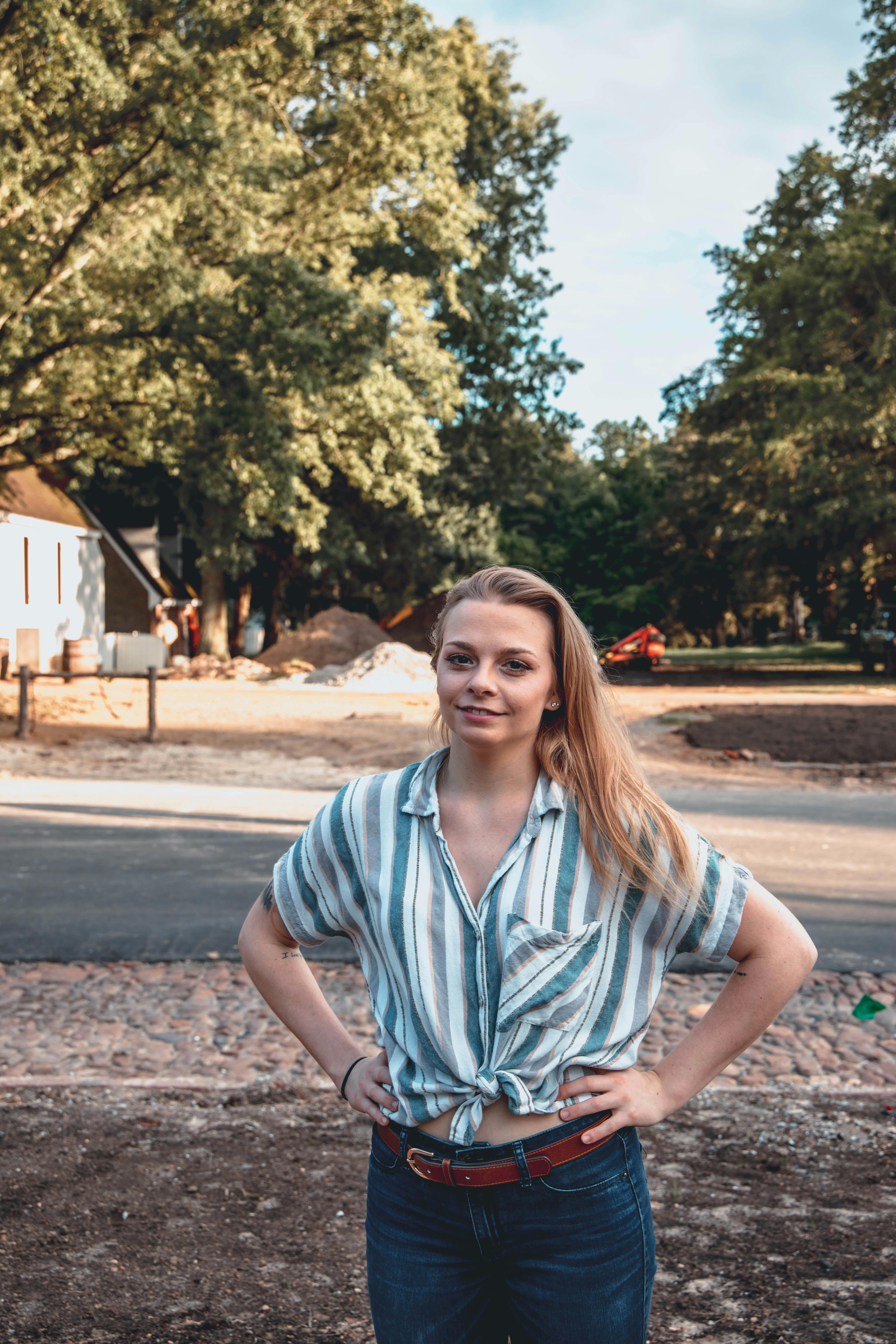 A confident woman with blond hair poses outdoors in a striped shirt, showcasing a warm summer vibe.