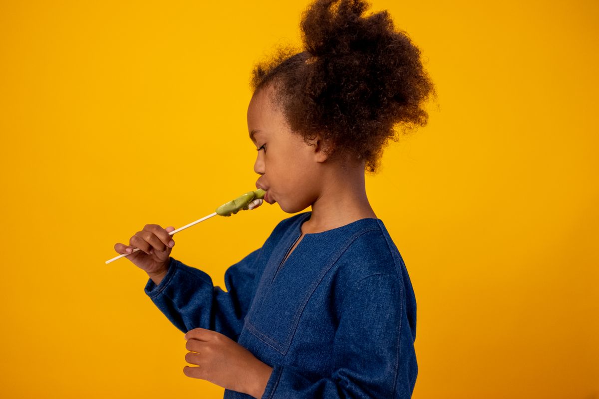 Adorable child with afro hair in blue dress enjoying a lollipop against a vibrant yellow background.