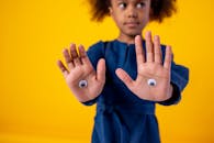 一名身穿蓝色连衣裙的年轻女孩，在亮黄色背景下伸出了手掌，手掌上装饰着粘稠的眼睛