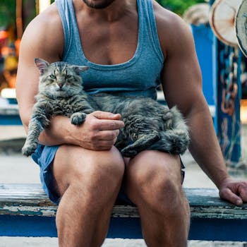 A muscular man in a tank top holding a relaxed cat outdoors on a bench.