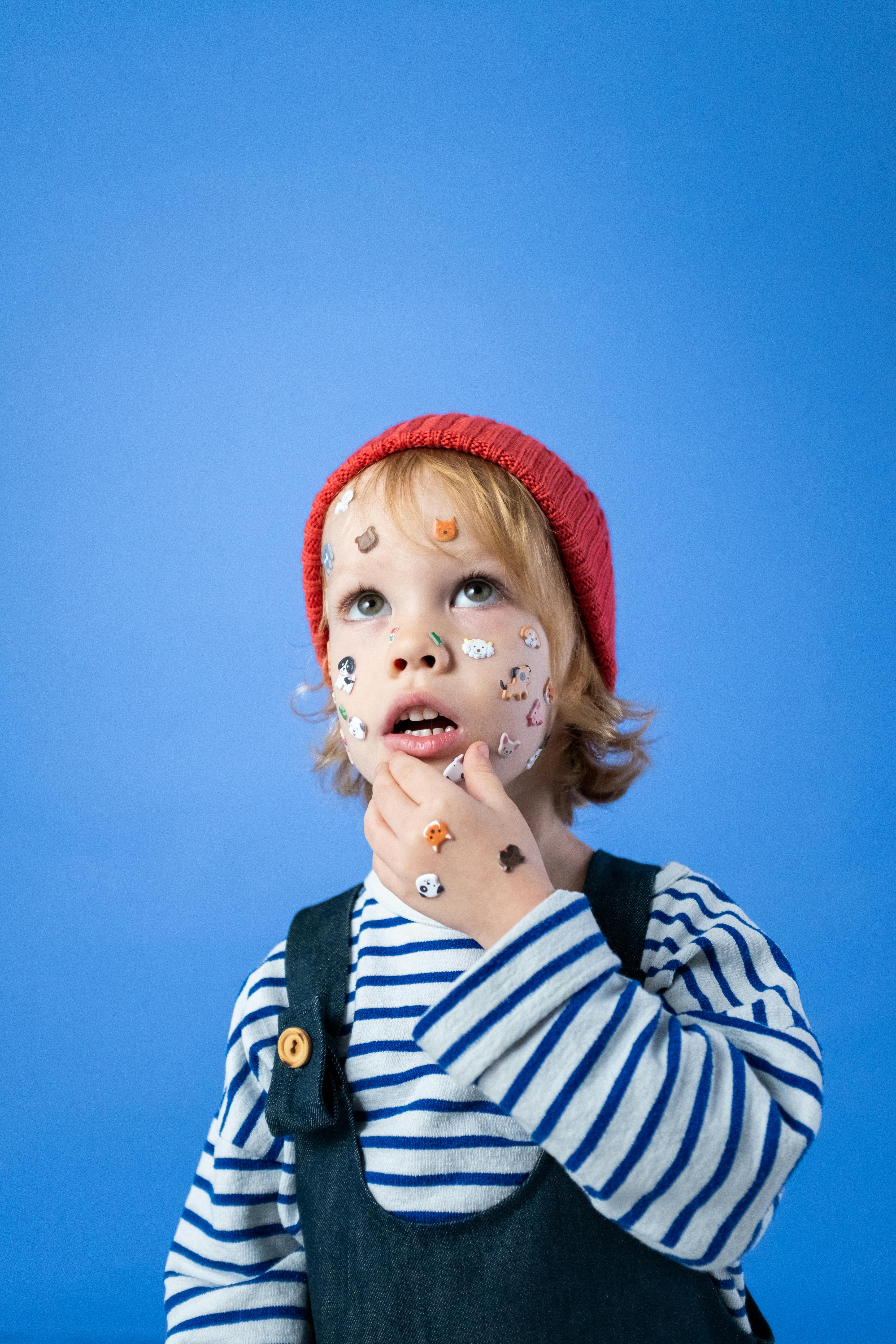 在蓝色工作室背景下，一个贴着脸贴、戴着红色帽子的可爱孩子显得若有所思