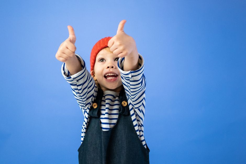 Happy child in striped shirt and red hat giving thumbs up, radiating joy against a blue background.