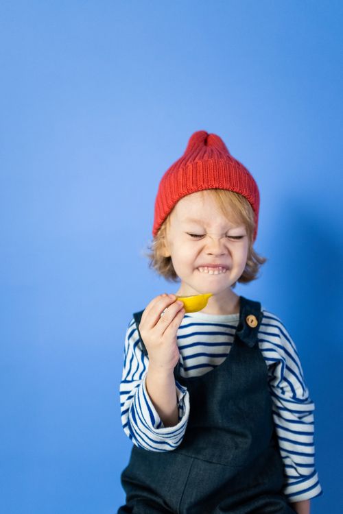 Free Cute child in striped outfit with red hat squints while trying a lemon slice in a colorful studio setting. Stock Photo