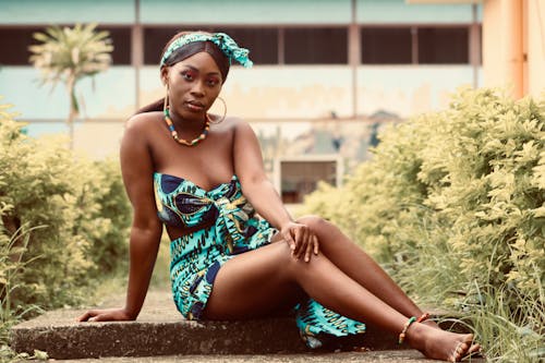 Full length gorgeous calm African American female in trendy summer dress sitting gracefully on stone ground in tropical garden and looking at camera