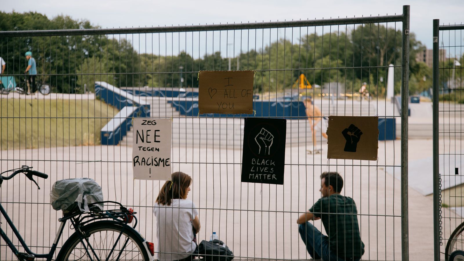 Back view of young cyclists sitting in skate park behind metal fence with signs against police brutality