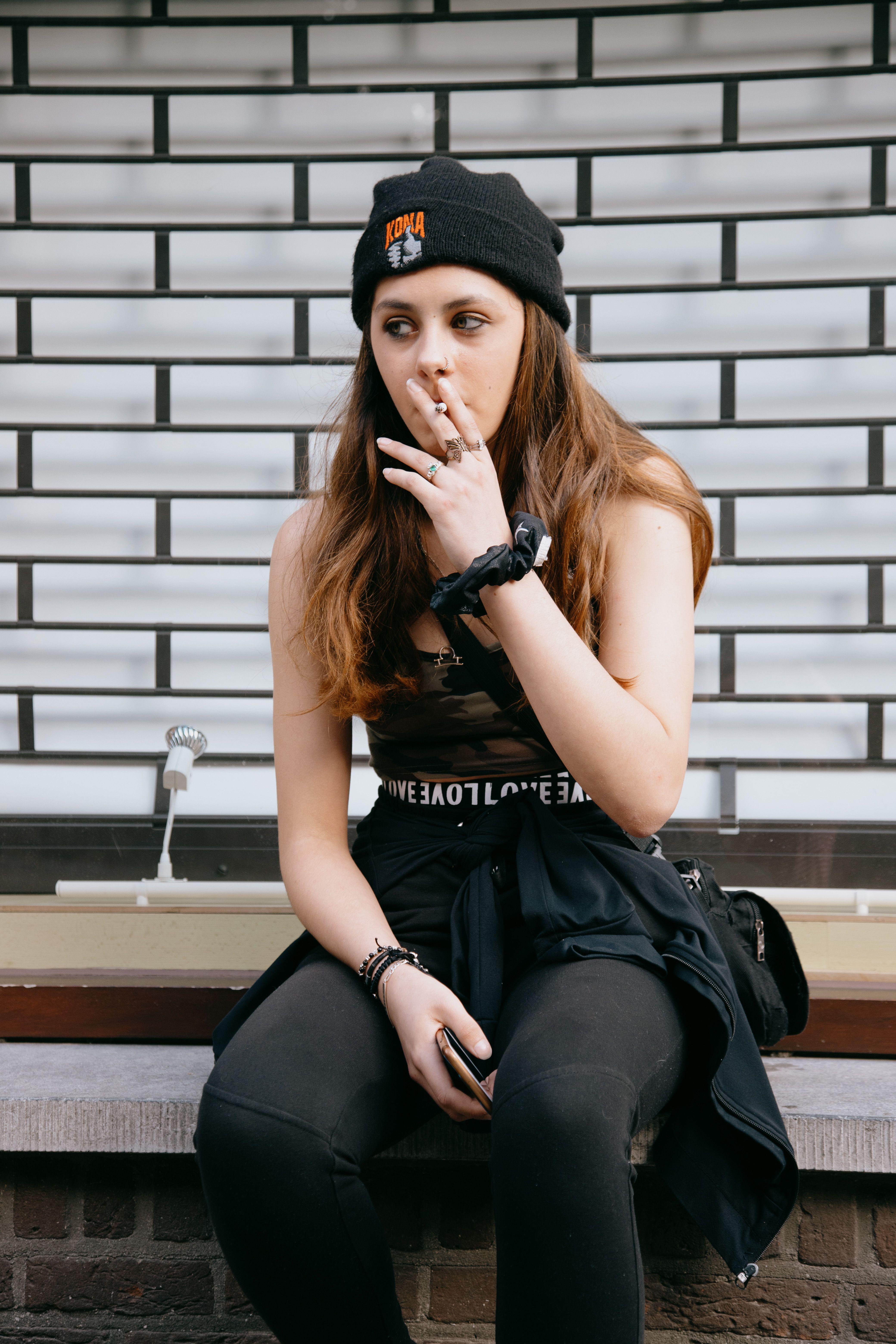 Female in trendy outfit sitting on stone border and smoking cigarette while looking away
