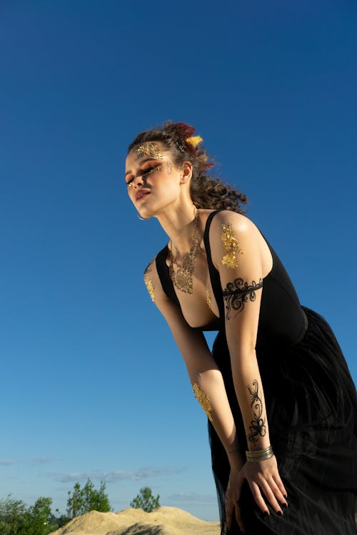 From below of beautiful young female with tattoos in black dress leaning over standing against blue cloudless sky