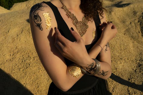 Crop anonymous female with tattoos and curly hair in black top standing with hands crossed in sand area