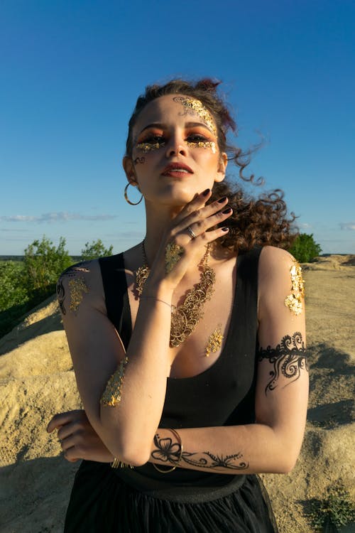 Young female with curly hair and tattoos and accessorizes standing on windy sandy terrain at sunny day