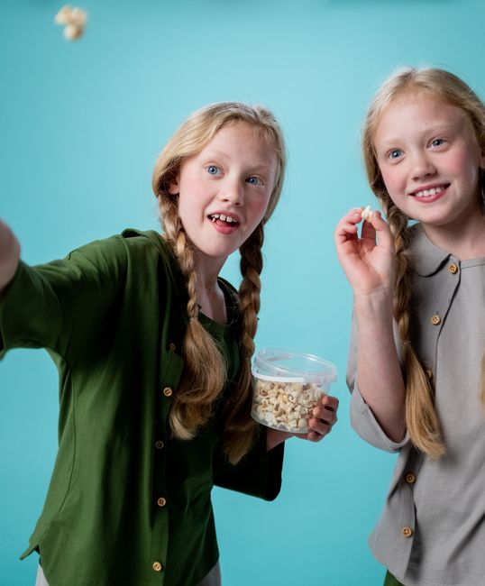 Happy twin sisters with braids enjoying popcorn against a vivid turquoise backdrop.