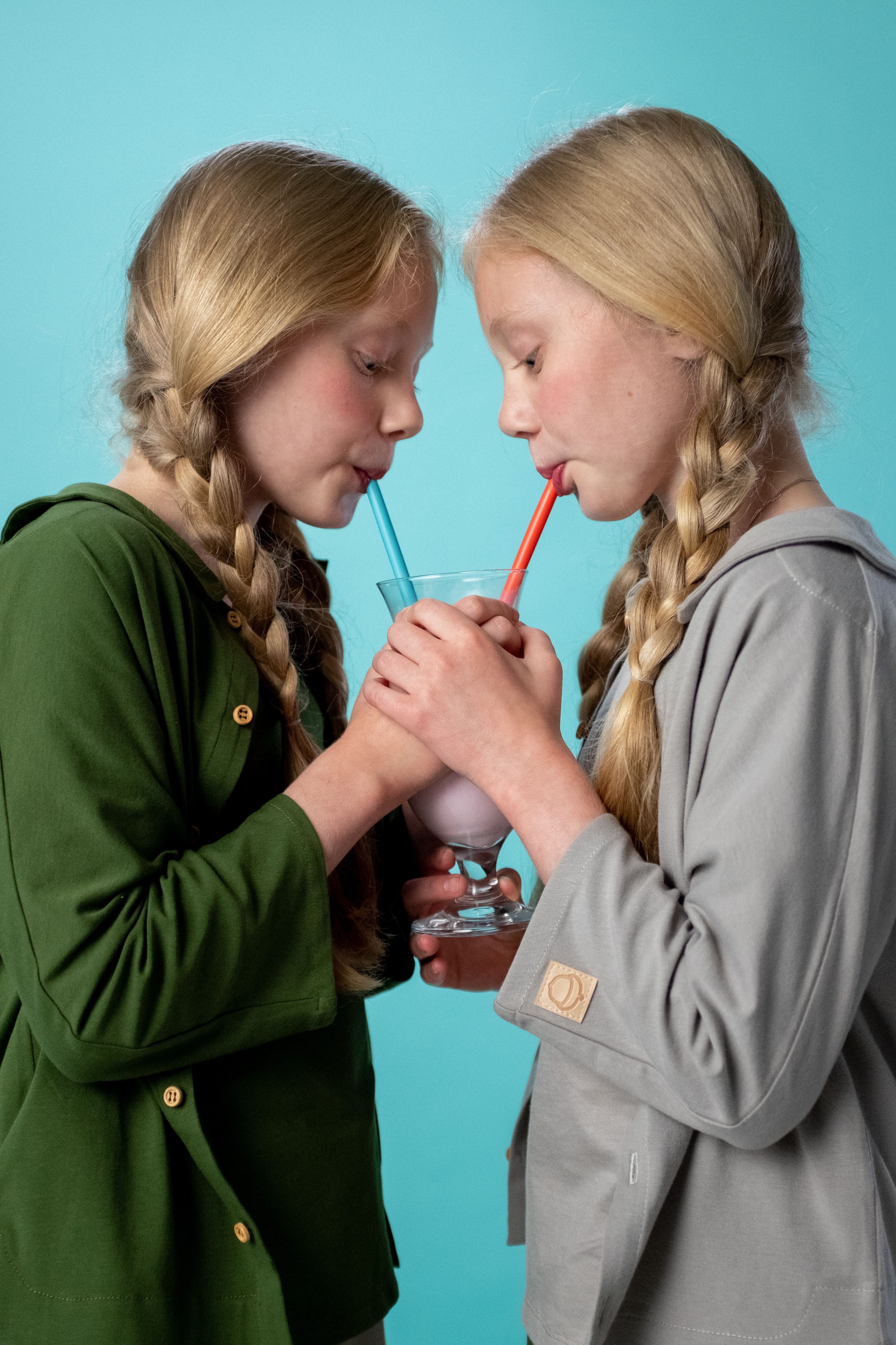 Two blonde twin girls enjoying a milkshake together. Studio setting with a turquoise background.