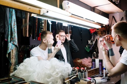 Free A man applies makeup in a dressing room mirror, surrounded by wigs and cosmetics. Stock Photo