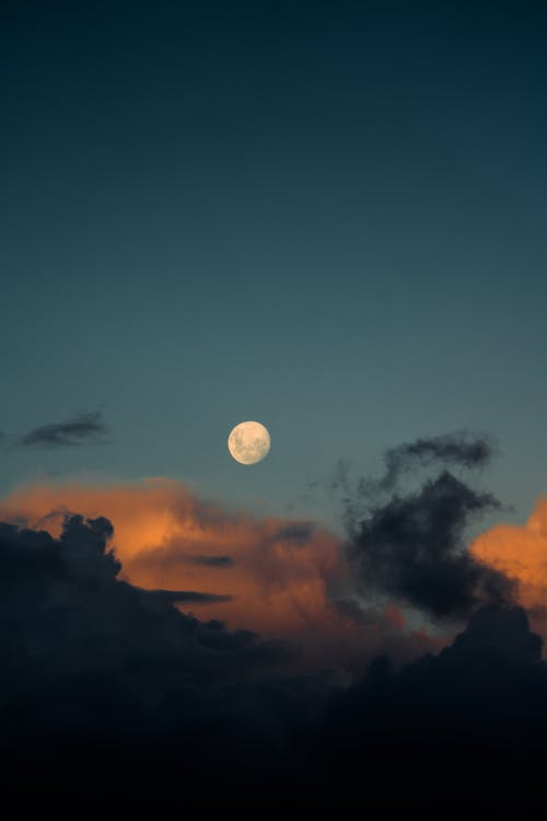 Glowing white moon with spots among cumulus gray cloudy in blue sky in evening