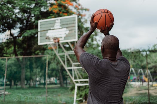 An adult man playing basketball outdoors, aiming for a shot in a lush park setting.