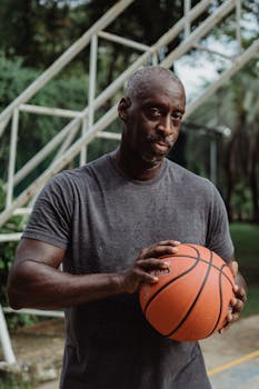 Pensive basketball player holding basketball on an outdoor court, expressing a serious demeanor.