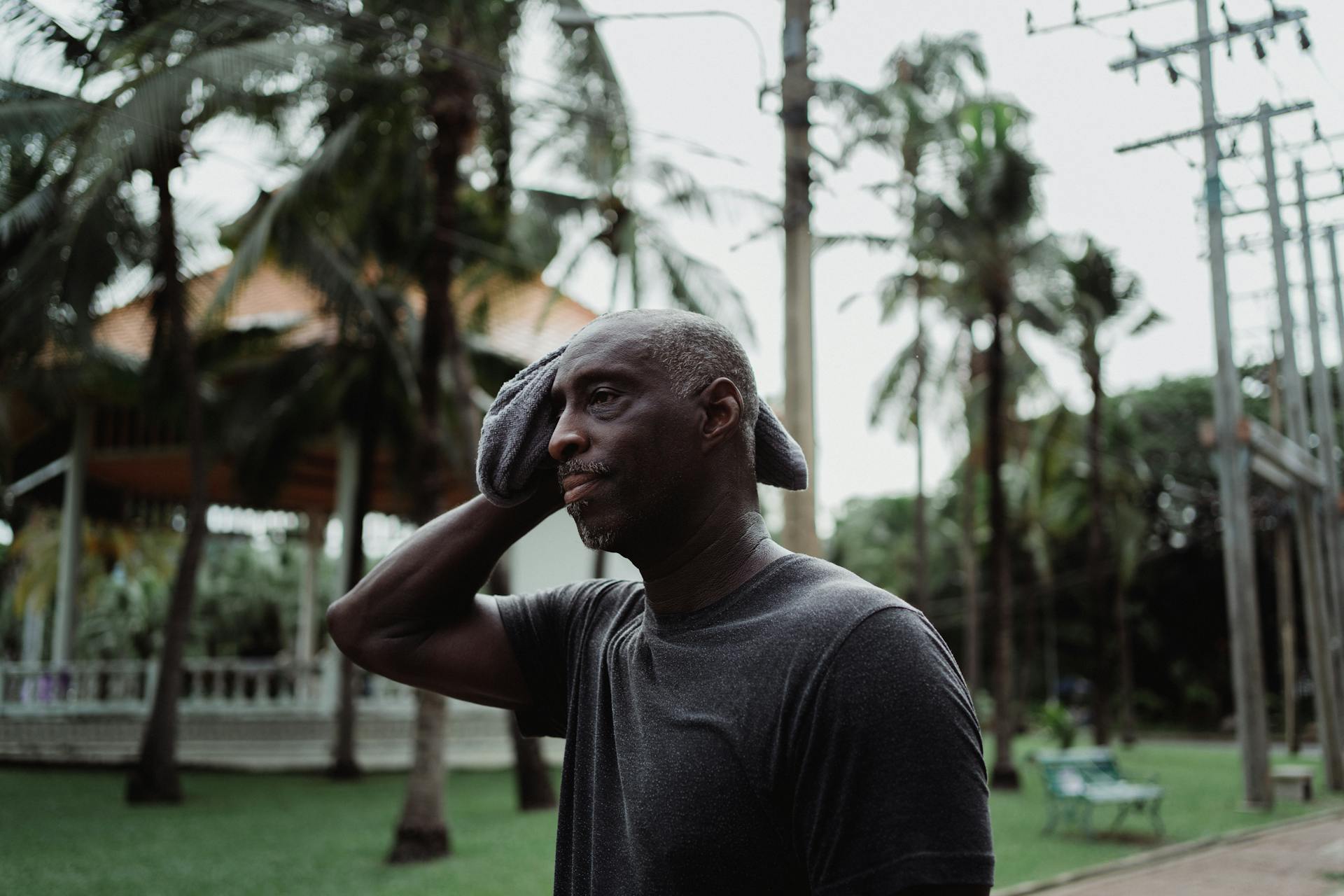 A man wiping sweat with a face towel outdoors in a tropical environment, appearing thoughtful and serious.