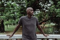 一名身穿灰色衬衫的男子，正站在桥上，对着郁郁葱葱的公园里那片宁静的湖水沉思