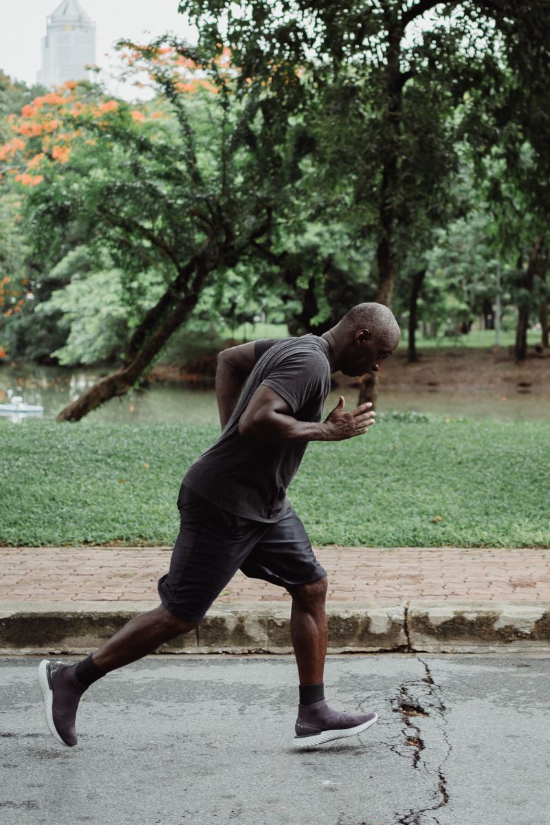 A Black man in a grey t-shirt and shorts running on a path through a lush green park, shown in mid-stride profile.