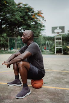 A man in sportswear sitting on a basketball, taking a break at an outdoor court.