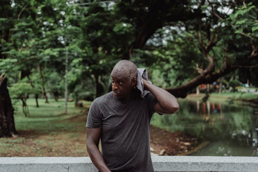 A man wipes his sweat with a towel while standing pensively outdoors in a lush park environment.