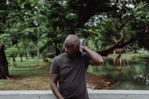 Free A man wipes his sweat with a towel while standing pensively outdoors in a lush park environment. Stock Photo