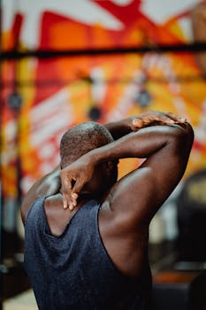 Back view of a muscular black man stretching arms indoors in a vibrant gym setting.