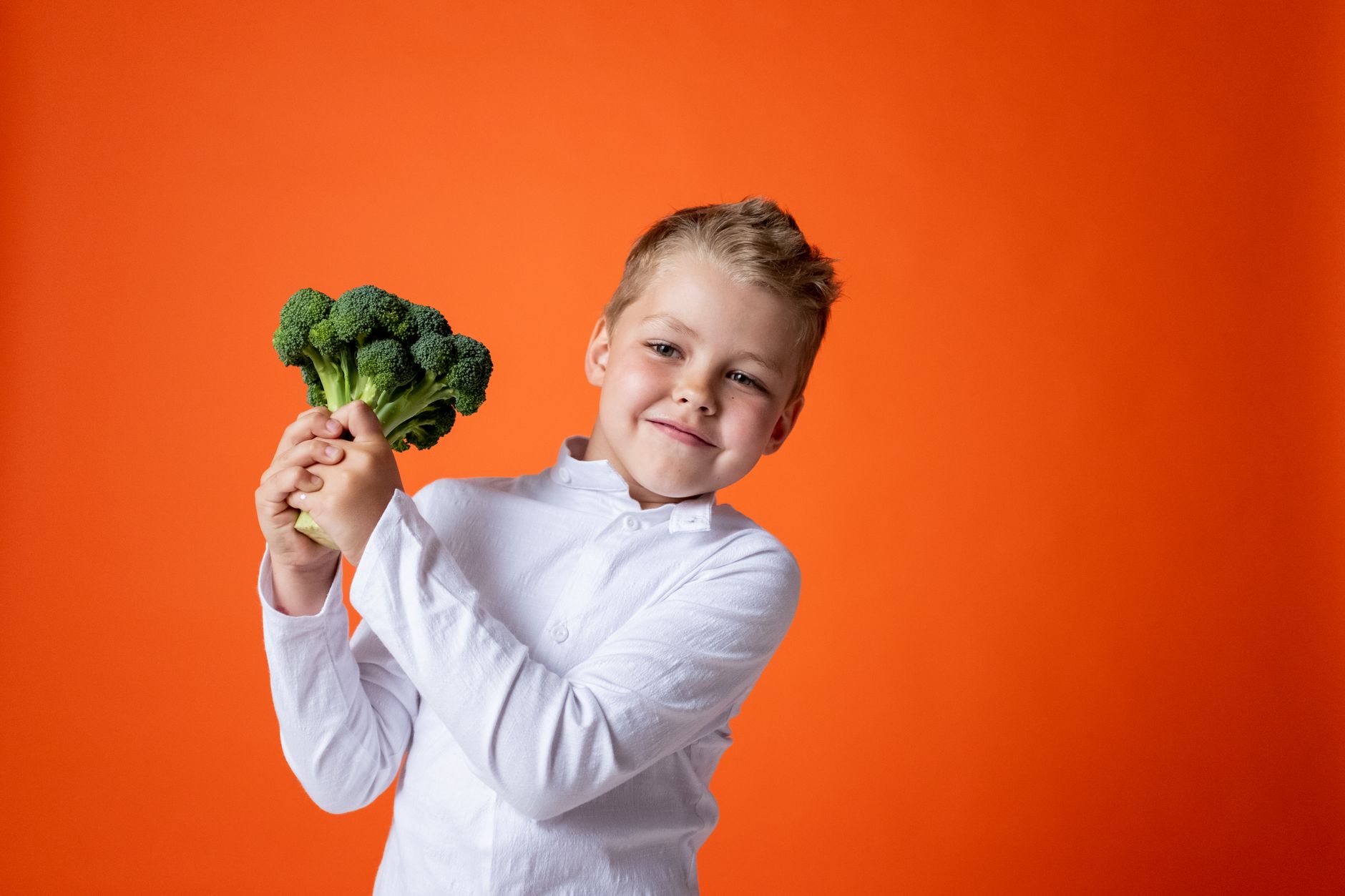 Cheerful young boy holding broccoli with a bright orange background.
