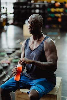 Fit African American man taking a break, holding a protein shake in a modern gym setting.