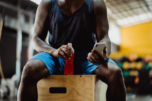 Free Athlete seated on box holding a smartphone and energy drink, blurred gym background. Stock Photo