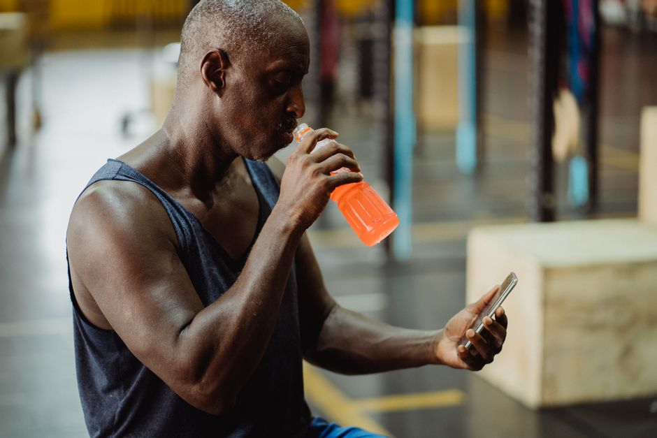 ジムでスマートフォンを使いながらスポーツドリンクを飲んでいるアフリカ系アメリカ人の男性。フィットネスと接続性のコンセプト。