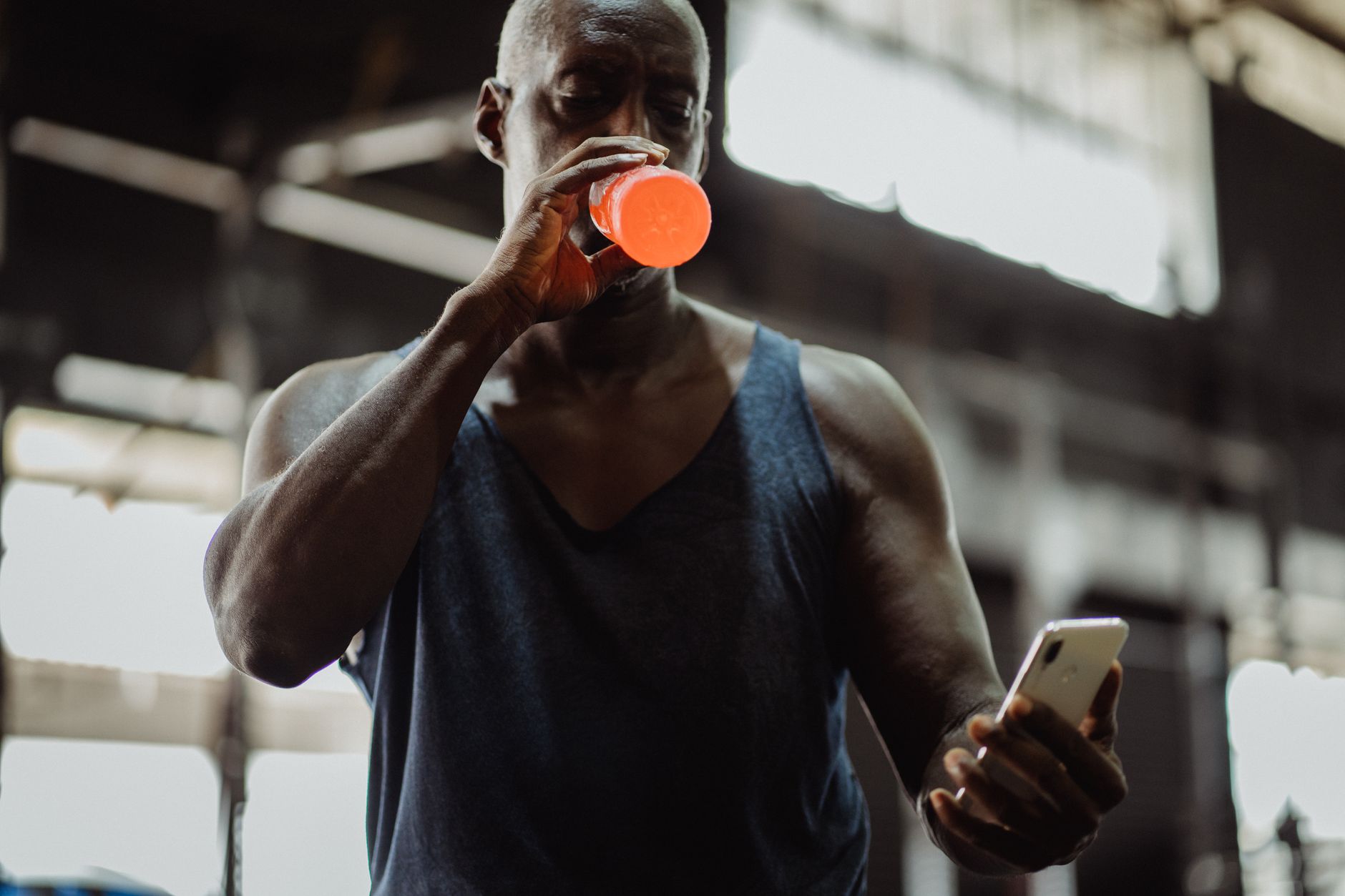 African American man drinking a sports drink while checking his smartphone in a gym setting.