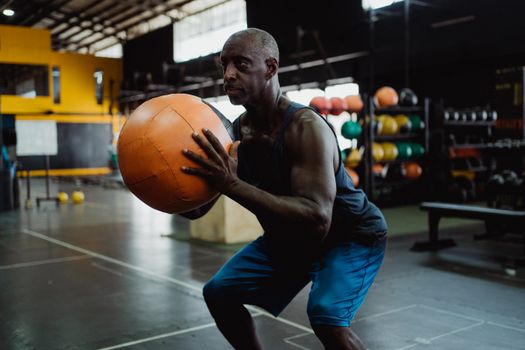 Focused mature man in gym squatting with a medicine ball during a workout.