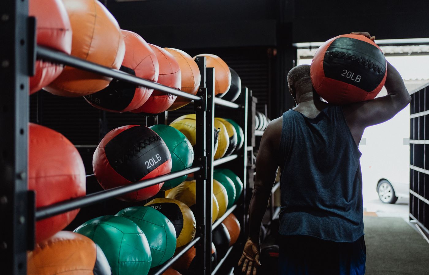 Back view of a man carrying a 20LB medicine ball in a gym with colorful balls on racks.