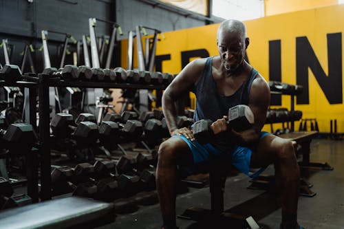 Free Man in Black Tank Top and Blue Shorts Sitting on Bench Lifting a Dumbbell Stock Photo