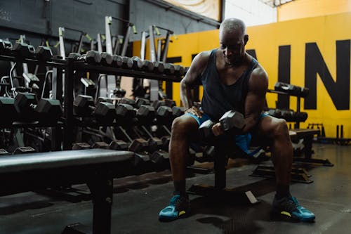 Man in Black Tank Top and Blue Shorts Sitting on Bench Lifting a Dumbbell