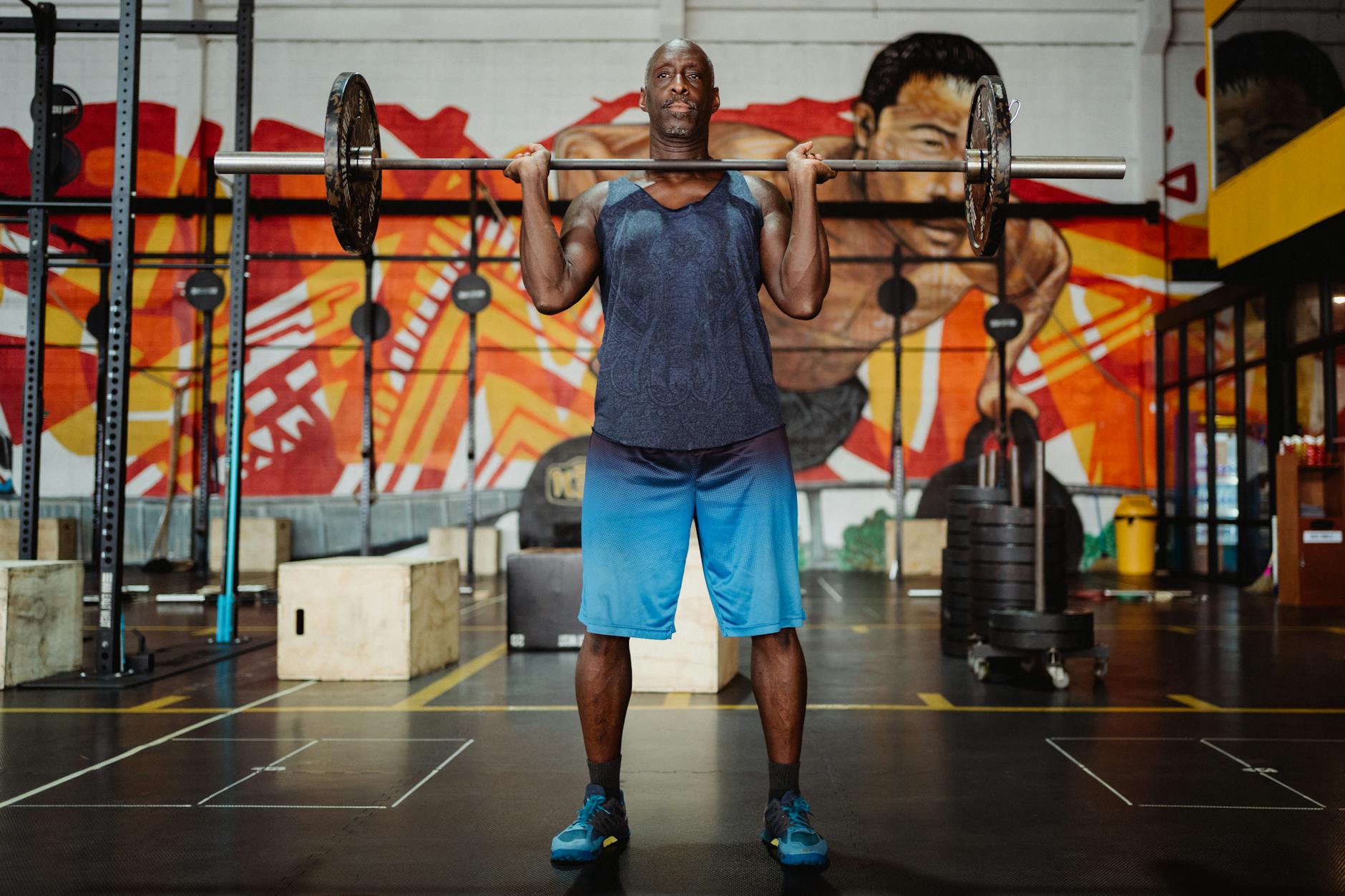 Strong man lifting a barbell inside a gym with colorful mural, emphasizing fitness and strength.