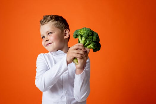 Cheerful young boy in white shirt holding broccoli against an orange studio backdrop.