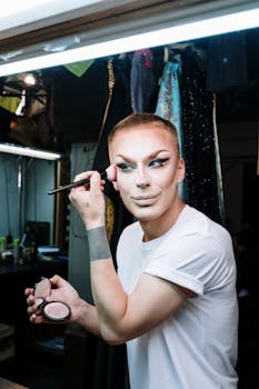 A drag artist applies makeup in a dressing room, emphasizing gender expression and transformation.