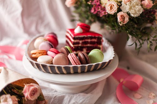 Stack of small round multi colored macaroons on saucer near cup of tea with piece of cake and bouquet of flowers in vase in bright room on blurred background