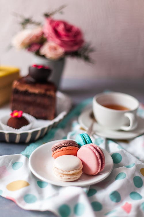 Overhead view of small round multi colored macaroons with cup of tea next to pink roses on table in bright cozy kitchen