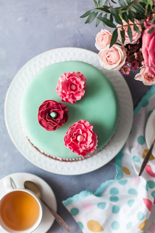 Overhead view of round turquoise cake covered with edible flowers between cup of tea and bouquet of roses with dark green leaves on table in daylight