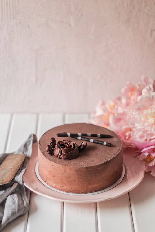 Round appetizing chocolate cake near bouquet of pink flowers on white table next to pink wall with uneved shabby surface