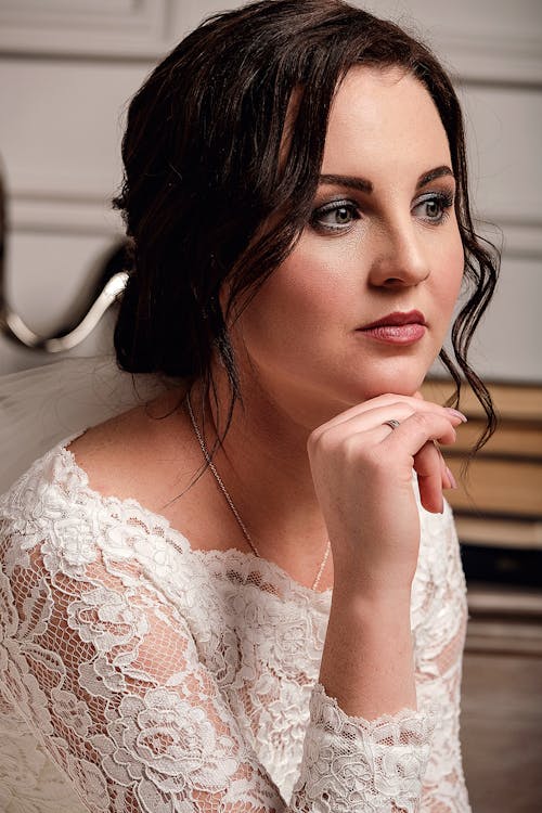 Pensive young bride in elegant white lace dress sitting in room with hand at chin and looking away before wedding ceremony
