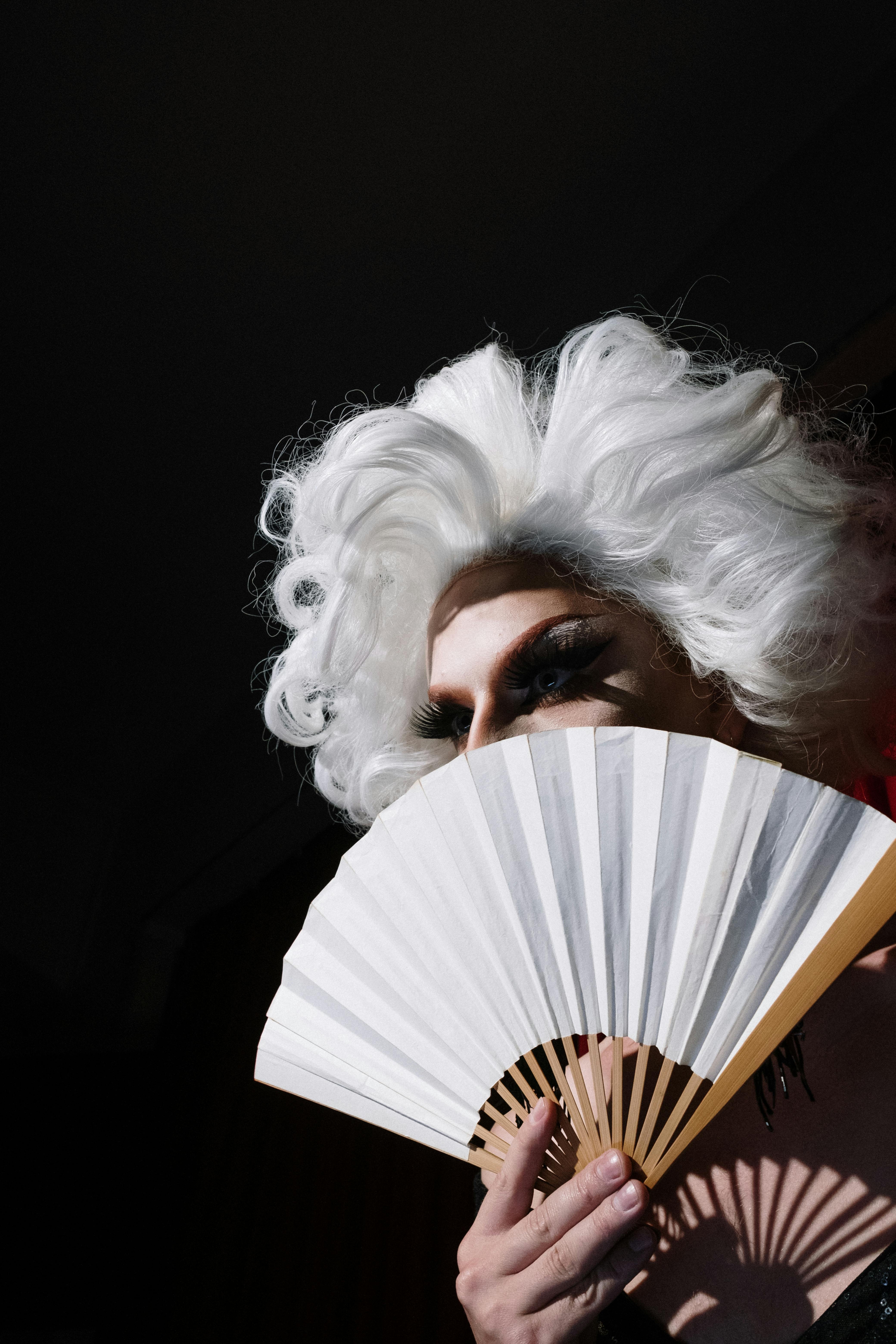 Free Elegant drag queen with striking white wig holding a fan under spotlight at night. Stock Photo