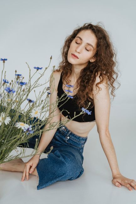 A serene woman with curly hair sits next to wildflowers with eyes closed in a minimalist studio setting.
