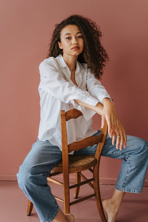 Woman in White Long Sleeves and Blue Denim Jeans Sitting on Wooden Chair
