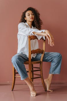 这幅肖像休闲而时尚，描绘了一位坐在木椅上的女人，背景朴素
