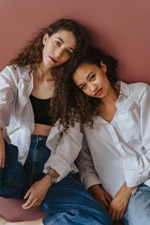 Free Two stylish women with curly hair sitting against a pink background in casual attire. Stock Photo